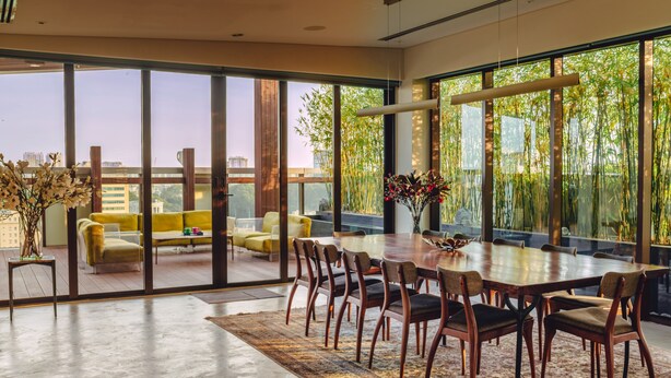 A spacious dining area with a large table and chairs, large windows with a view of greenery, and a yellow sofa in the background. - SILA Urban Living (Ho Chi Minh City)