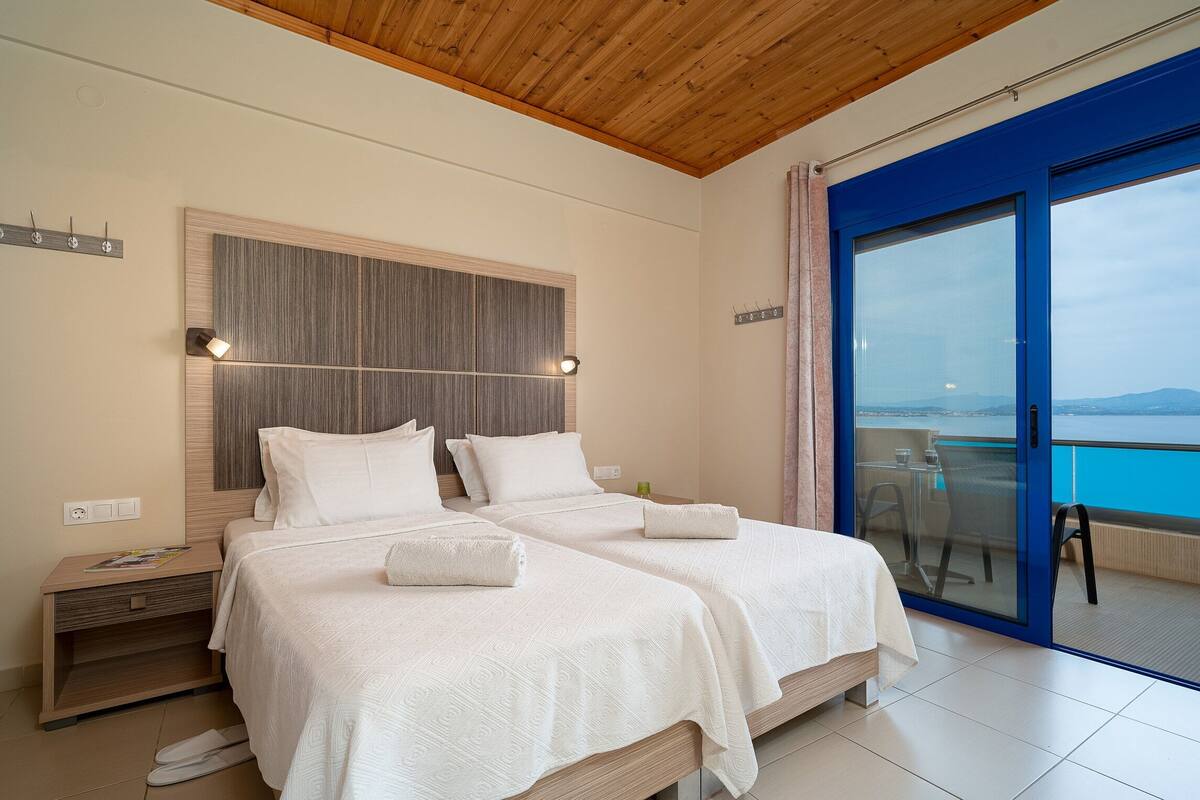 A hotel room with two beds, a wooden ceiling, a large window with a view, and a balcony with a table and chairs.