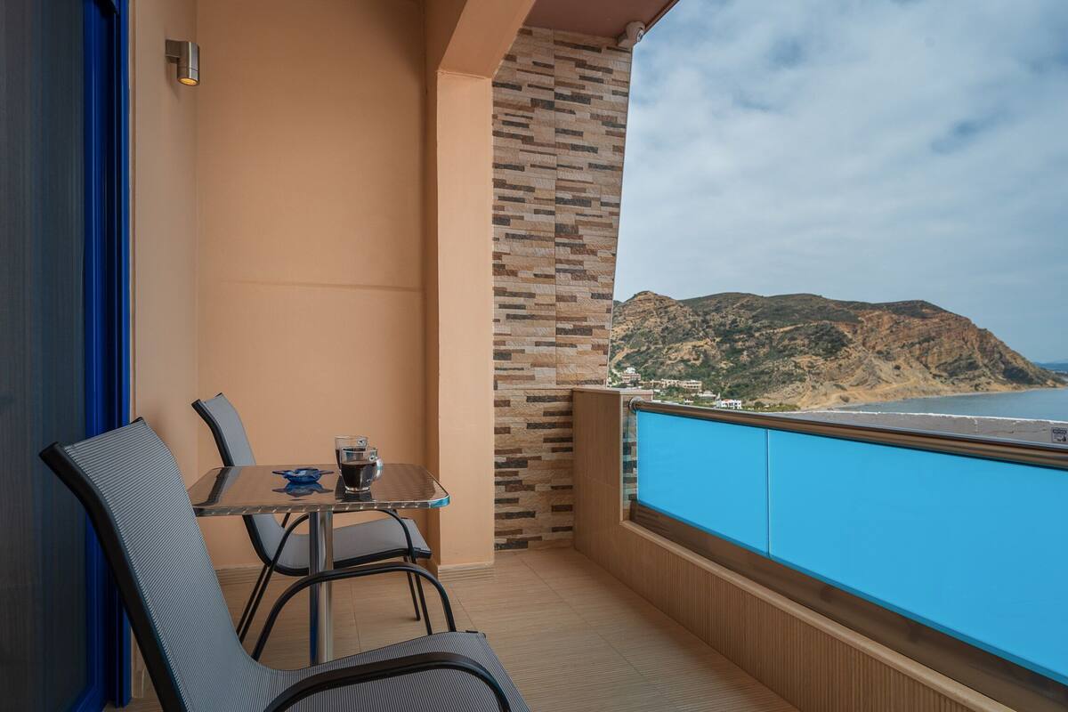 A balcony with a table and chairs, a view of a mountainous coastline, and a patterned wall.