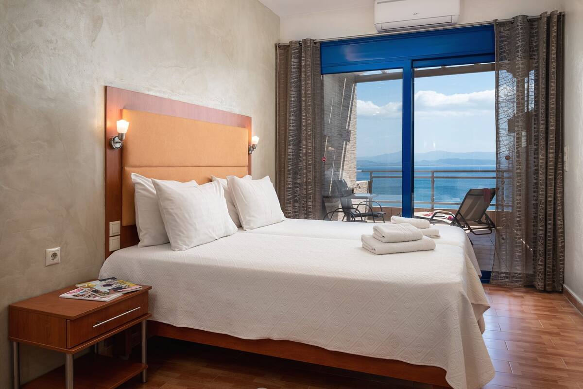 A double bed with a headboard, a bedside table with magazines, and a view of the sea through a large window with curtains.