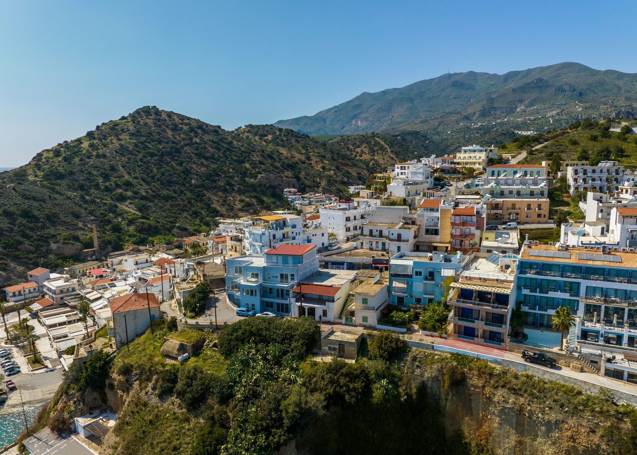 A coastal town with buildings built on a hillside, featuring a mix of modern and traditional architecture, and a clear view of the sea and mountains in the background.