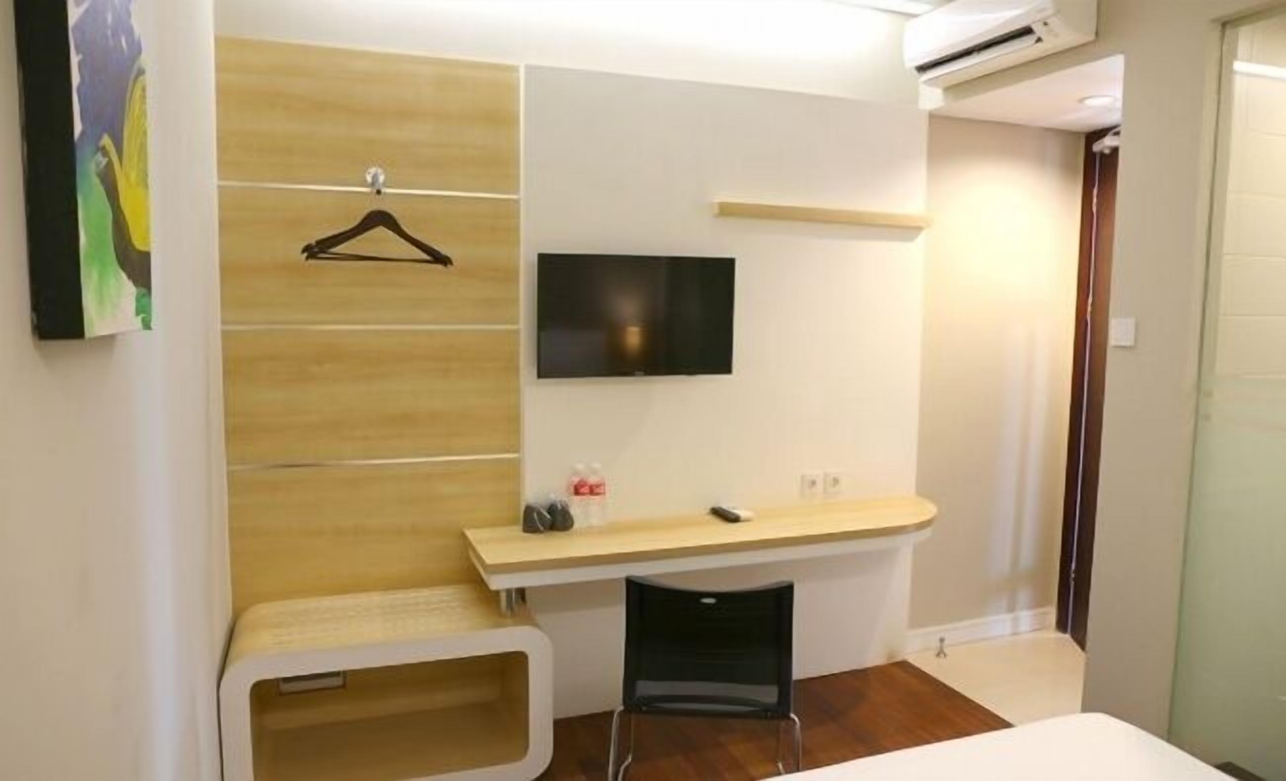 A hotel room with a wooden panel wall, a mounted TV, a small desk with a chair, and a bench.