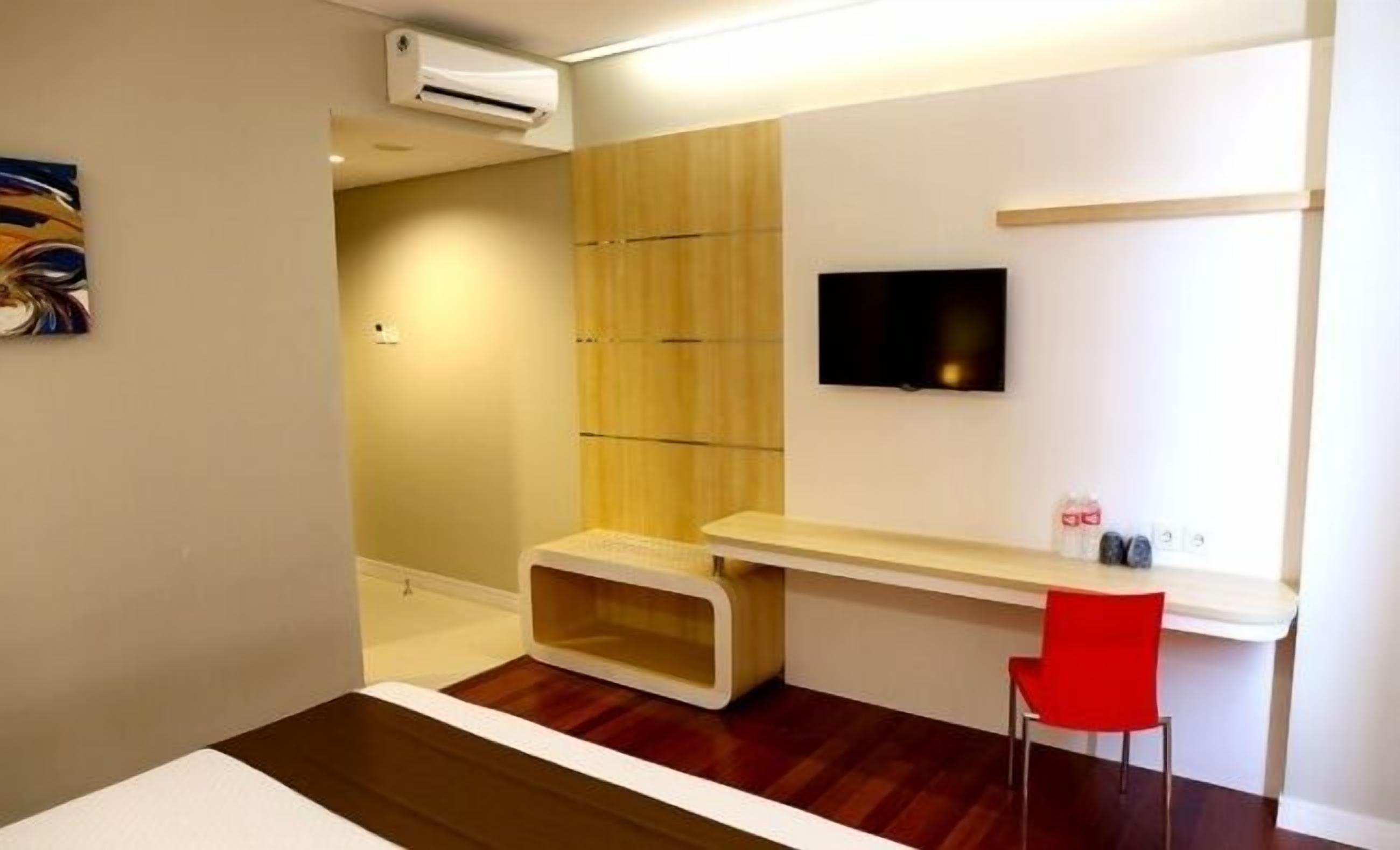 Habitación de hotel moderna con piso de madera, televisor en la pared, un escritorio pequeño y una silla roja.