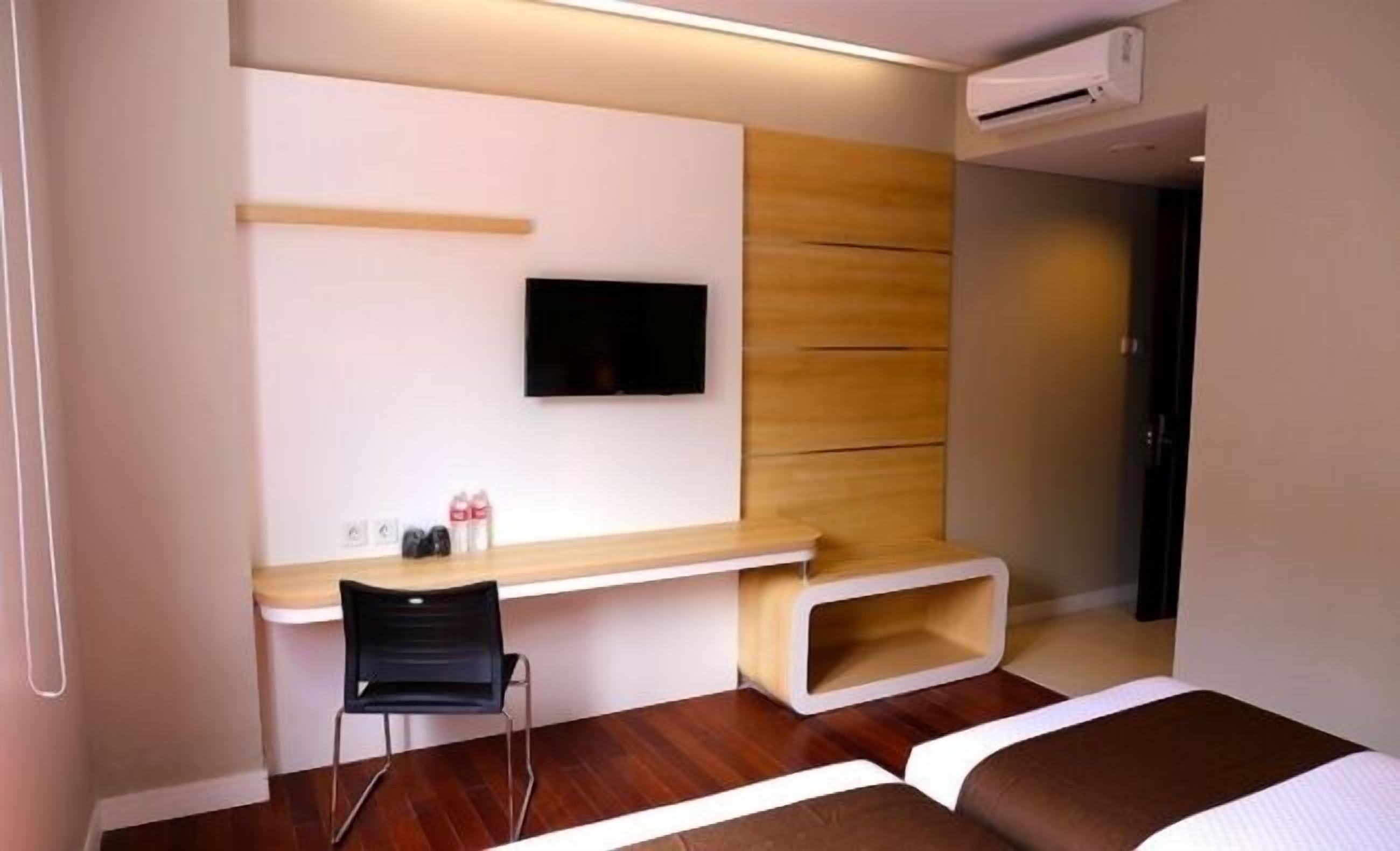 Una habitación de hotel moderna con un escritorio de madera, una silla negra, un televisor de pantalla plana montado en la pared y una cama con cabecera marrón.