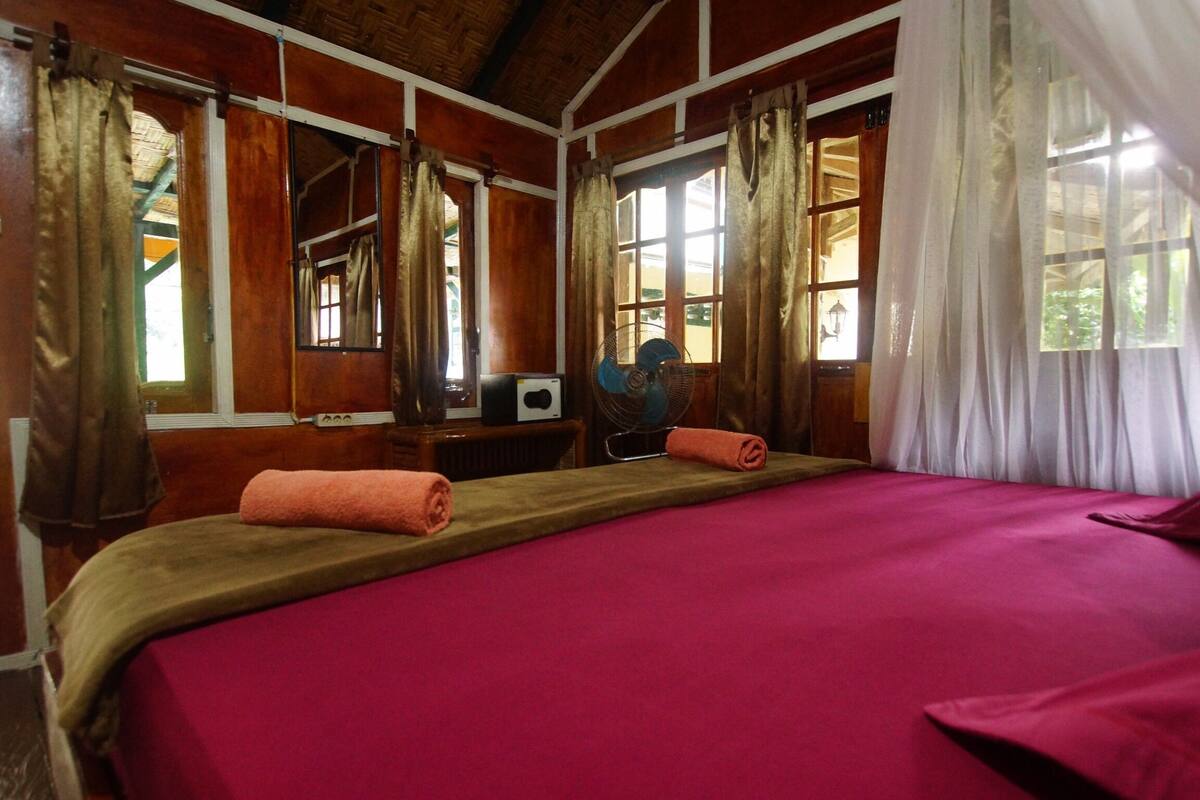 A room with a large bed, two windows with curtains, a desk with a fan, and a wooden ceiling.