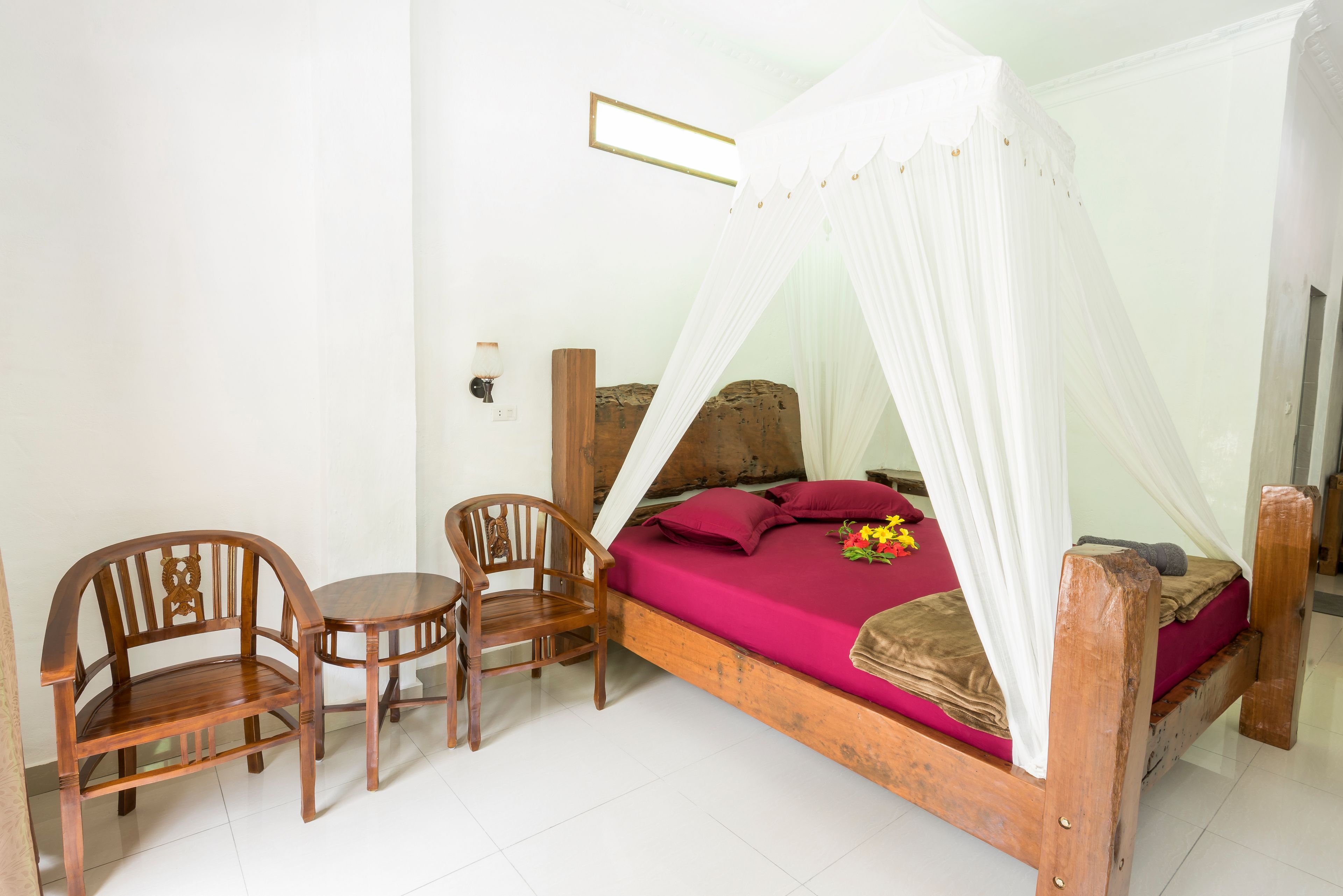 A wooden bed with a canopy, a wooden chair, and a small table in a room with white walls and a tiled floor.