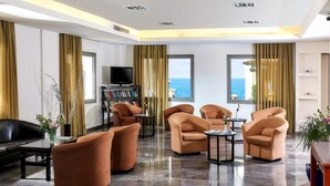 A modern hotel lobby with a large window offering a sea view, plush seating, a bookshelf, and a flat-screen TV. - Panormo Beach (Mylopotamos)