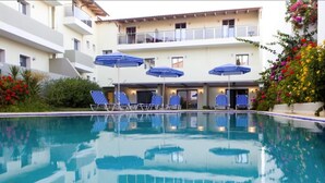 A pool area with lounge chairs and umbrellas, surrounded by a building with balconies and a garden with flowers. - Panormo Beach (Mylopotamos)