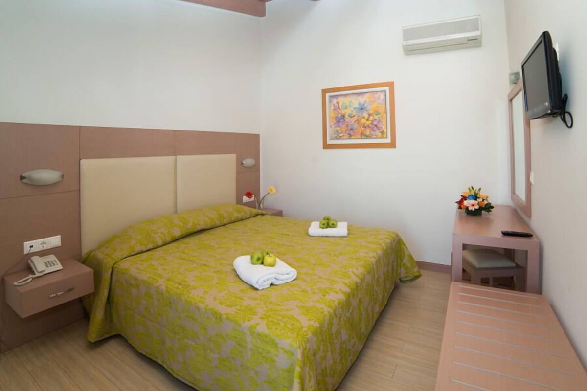 A hotel room with a bed, bedside table, a small desk with a chair, a television, and a painting on the wall.