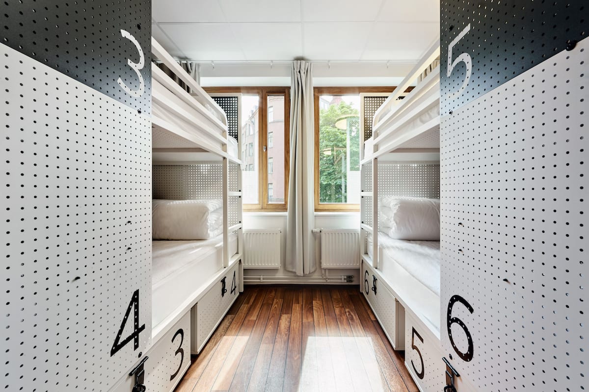 Una habitación de hotel moderna con literas numeradas del 3 al 6, suelo de madera y ventanas amplias con cortinas.