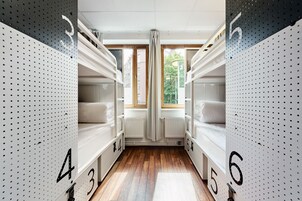 A modern hotel room with bunk beds numbered 3 to 6, wooden flooring, and large windows with curtains.
