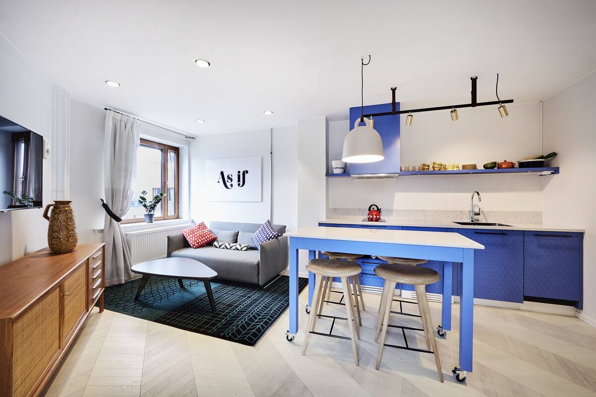 Una cocina moderna con encimera azul, un comedor con mesa y taburetes blancos, y una sala de estar con sofá gris y una mesita de centro.