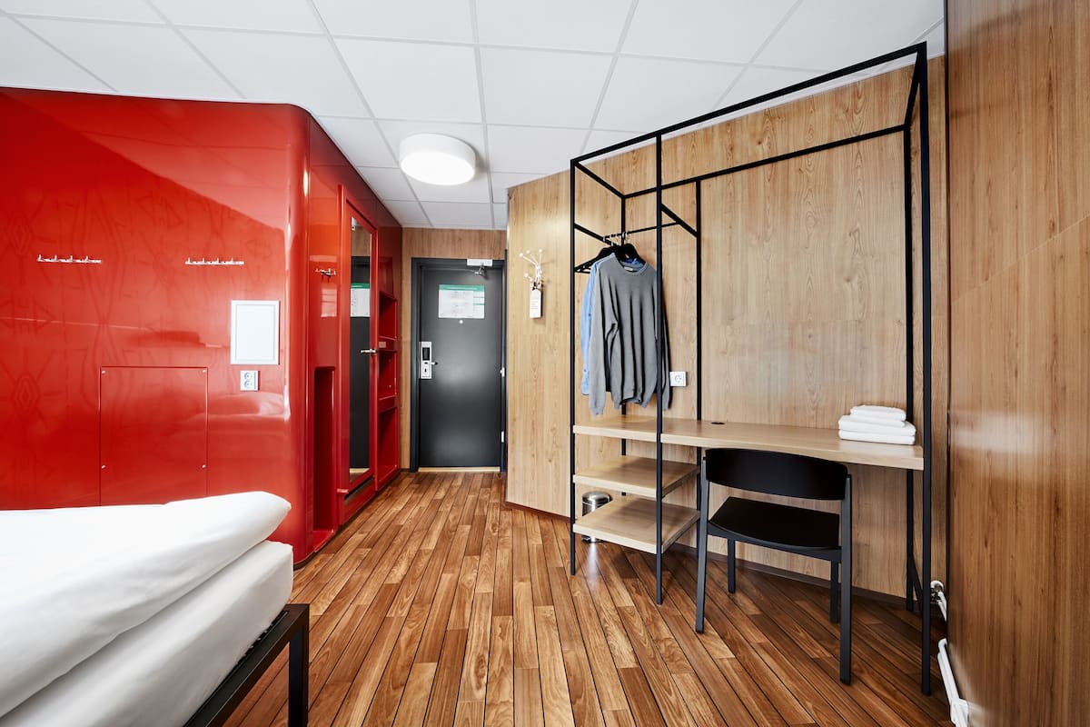 Una habitación de hotel moderna con suelo de madera, una pared de acento roja, una cama, un escritorio con una silla y un perchero.
