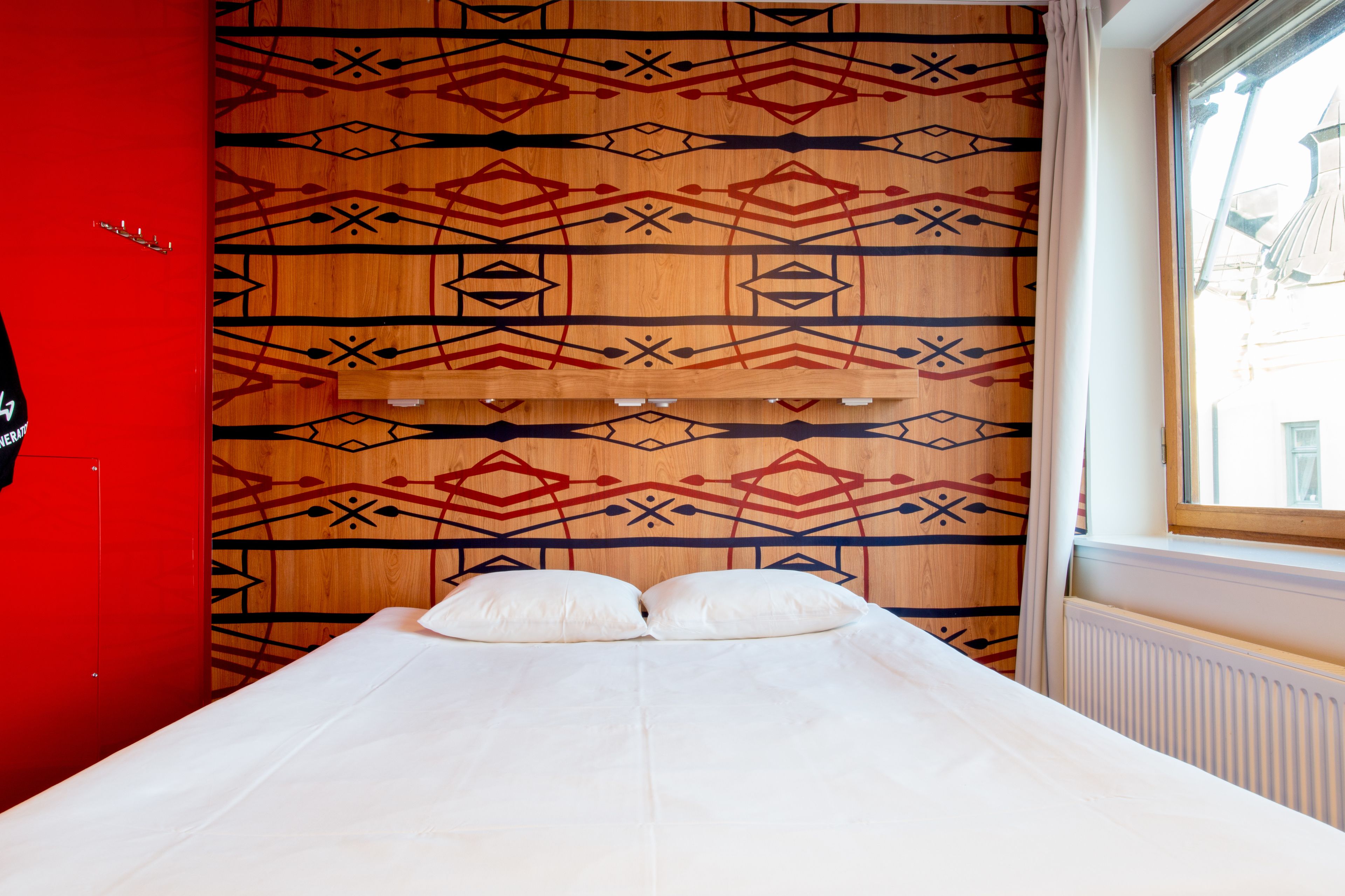A bed with a patterned headboard, a red wall, and a window with a view.