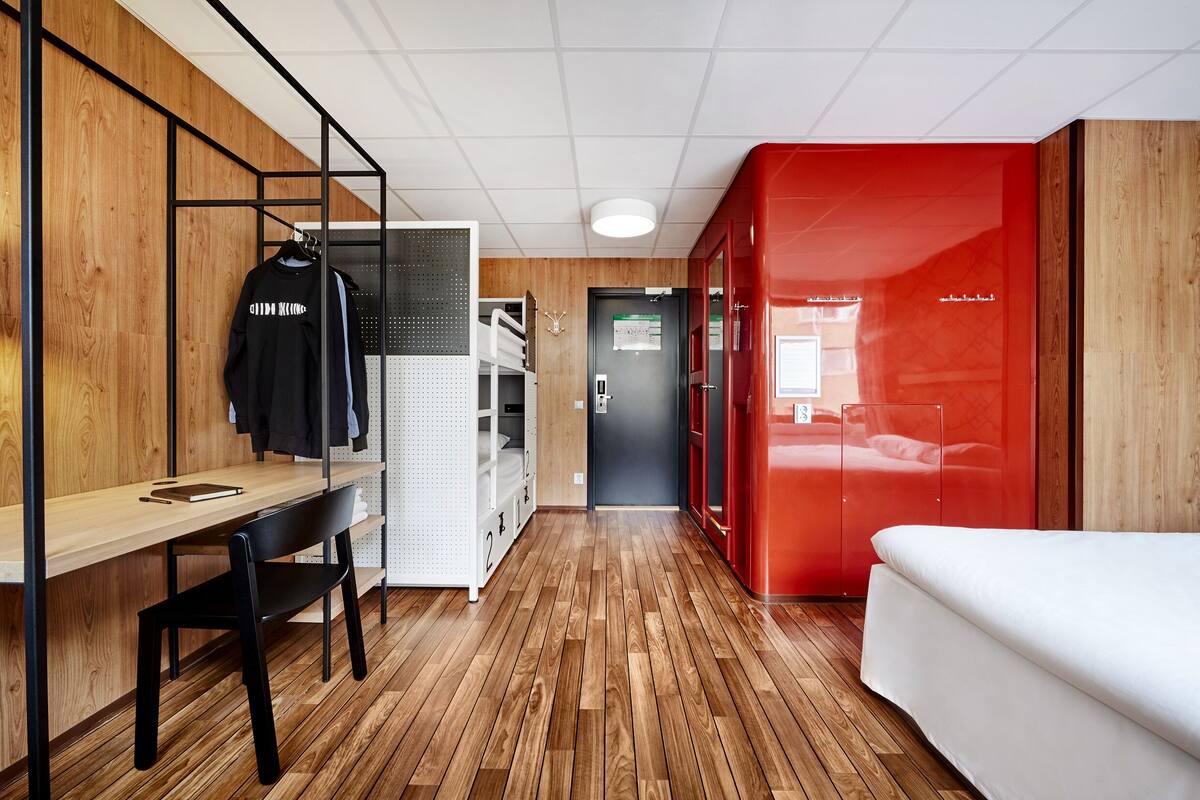 Una habitación de hotel moderna con piso de madera, un escritorio con silla, una cama y una pared de acento roja.