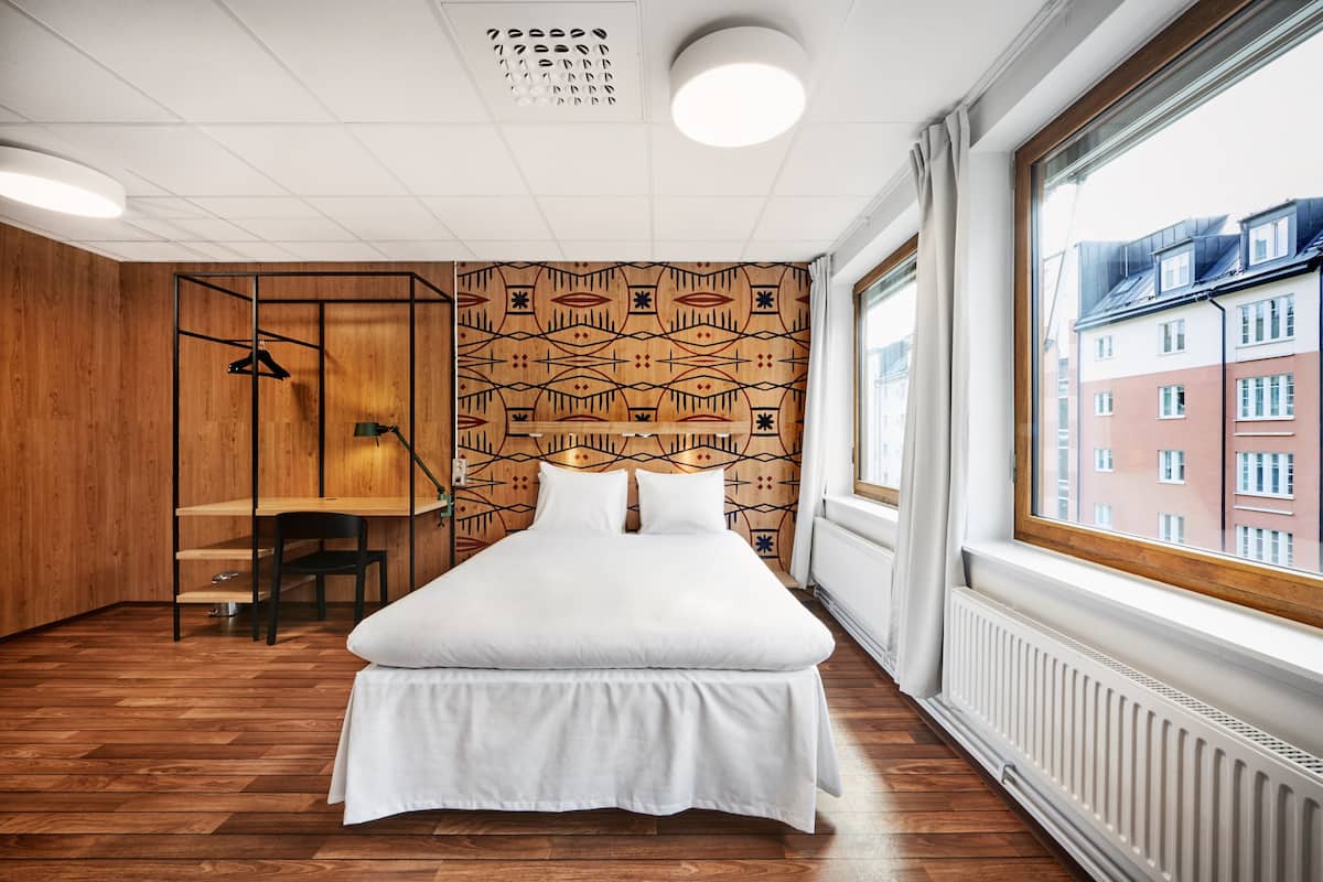 Una habitación de hotel moderna con suelo de madera, una cama grande con ropa blanca, un escritorio con silla y una pared con diseño.