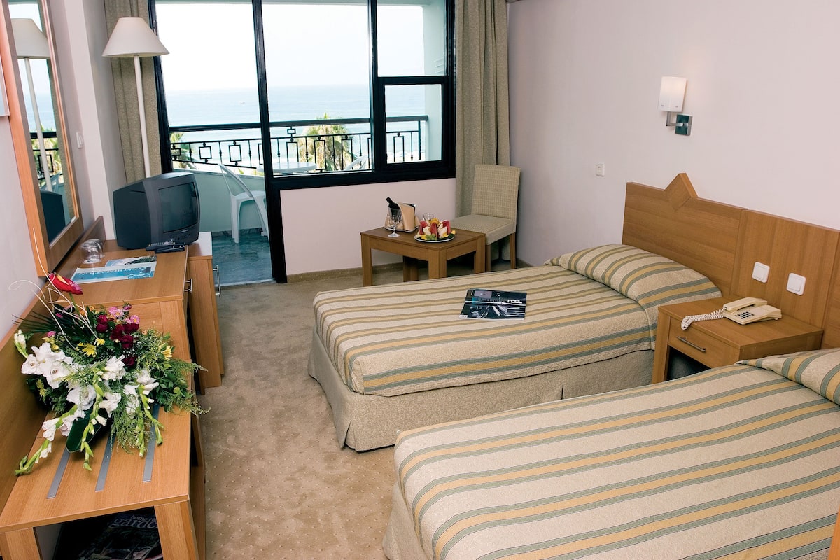 Habitación de hotel con dos camas, un escritorio con una lámpara, una mesita con un teléfono y un vaso, y vista al mar a través de amplias ventanas.
