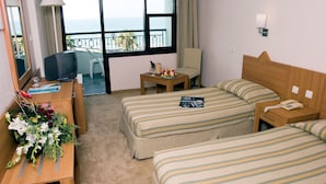 Quarto de hotel com duas camas, uma escrivaninha com luminária, uma mesinha com telefone e um copo, e vista para o mar através de grandes janelas.