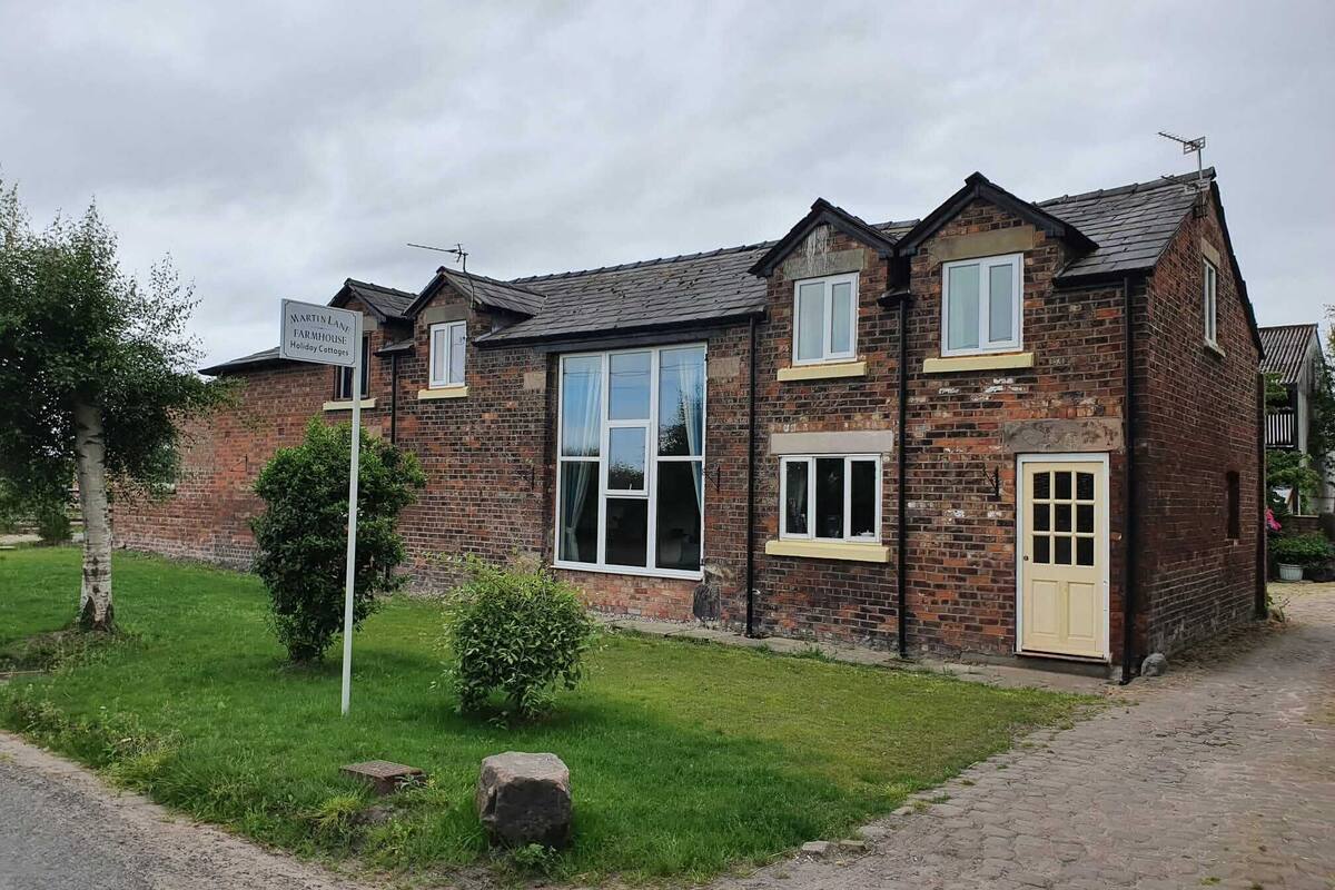 A two-story brick building with a large window and a door, surrounded by a grassy area and a paved path.