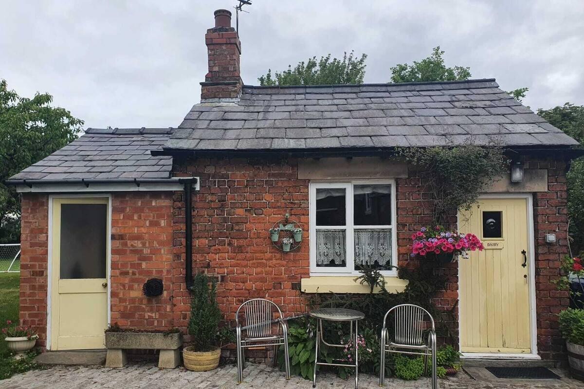 A small brick building with a sloped roof, a chimney, and a window with lace curtains. There are two metal chairs and a small table outside.