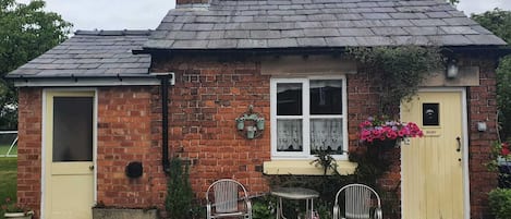 A small brick building with a sloped roof, a chimney, and a window with lace curtains. There are two metal chairs and a small table outside.