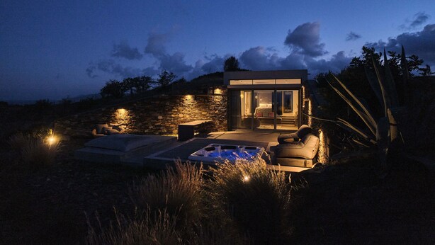 A modern outdoor hot tub area with a stone wall and seating, surrounded by tall grasses and a clear sky.