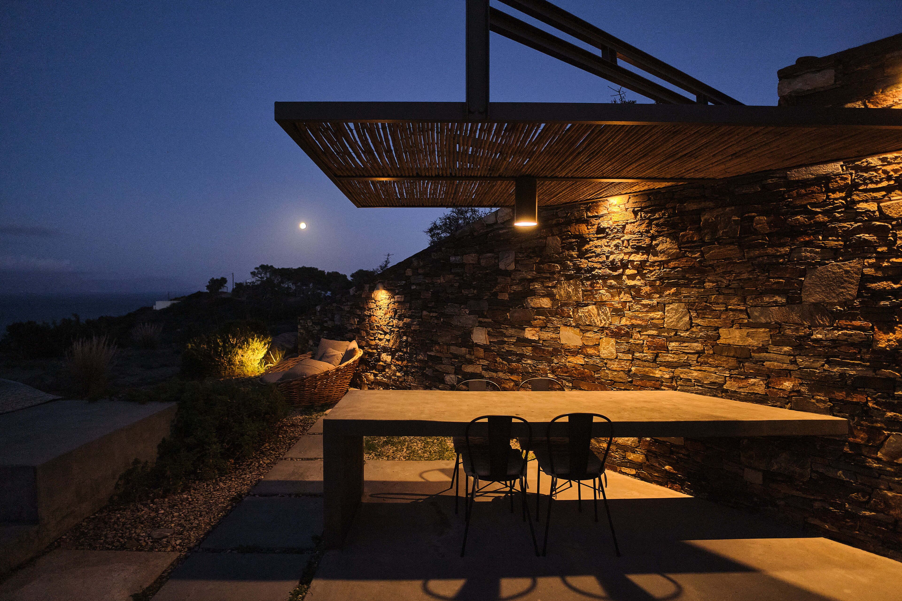 Een terras buiten, met een stenen muur, een tafel en stoelen, onder een houten pergola met verlichting.