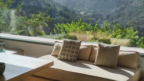 A modern room with a sofa and cushions, a glass table, and a view of a lush green landscape.