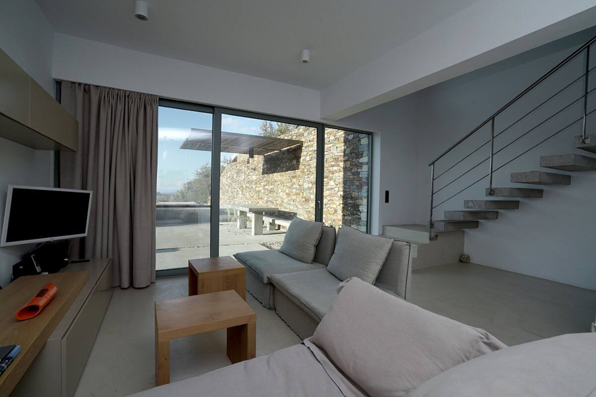 A modern living room with a sofa, a coffee table, and a TV. There is a staircase and a view of the outside.