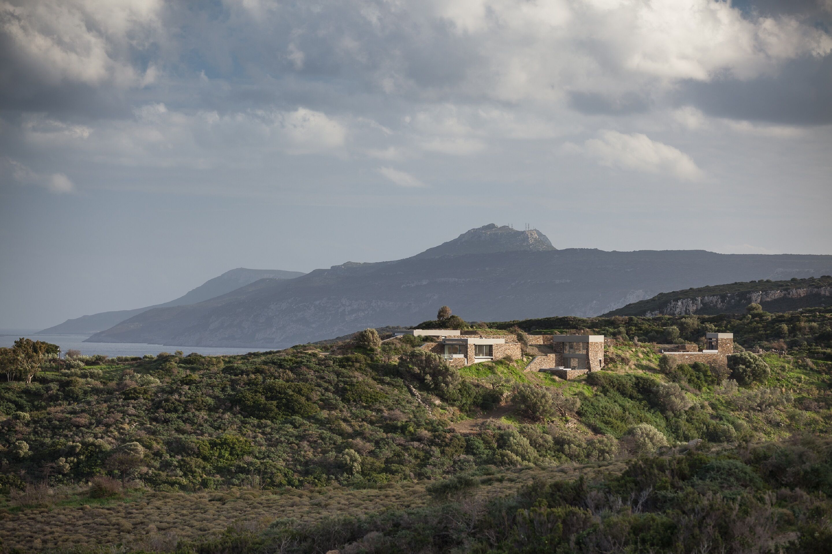 Una casa rústica y apartada en una ladera, con vistas al mar y a las montañas al fondo.