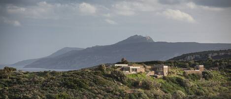 Una casa rústica y apartada en una ladera, con vistas al mar y a las montañas al fondo.
