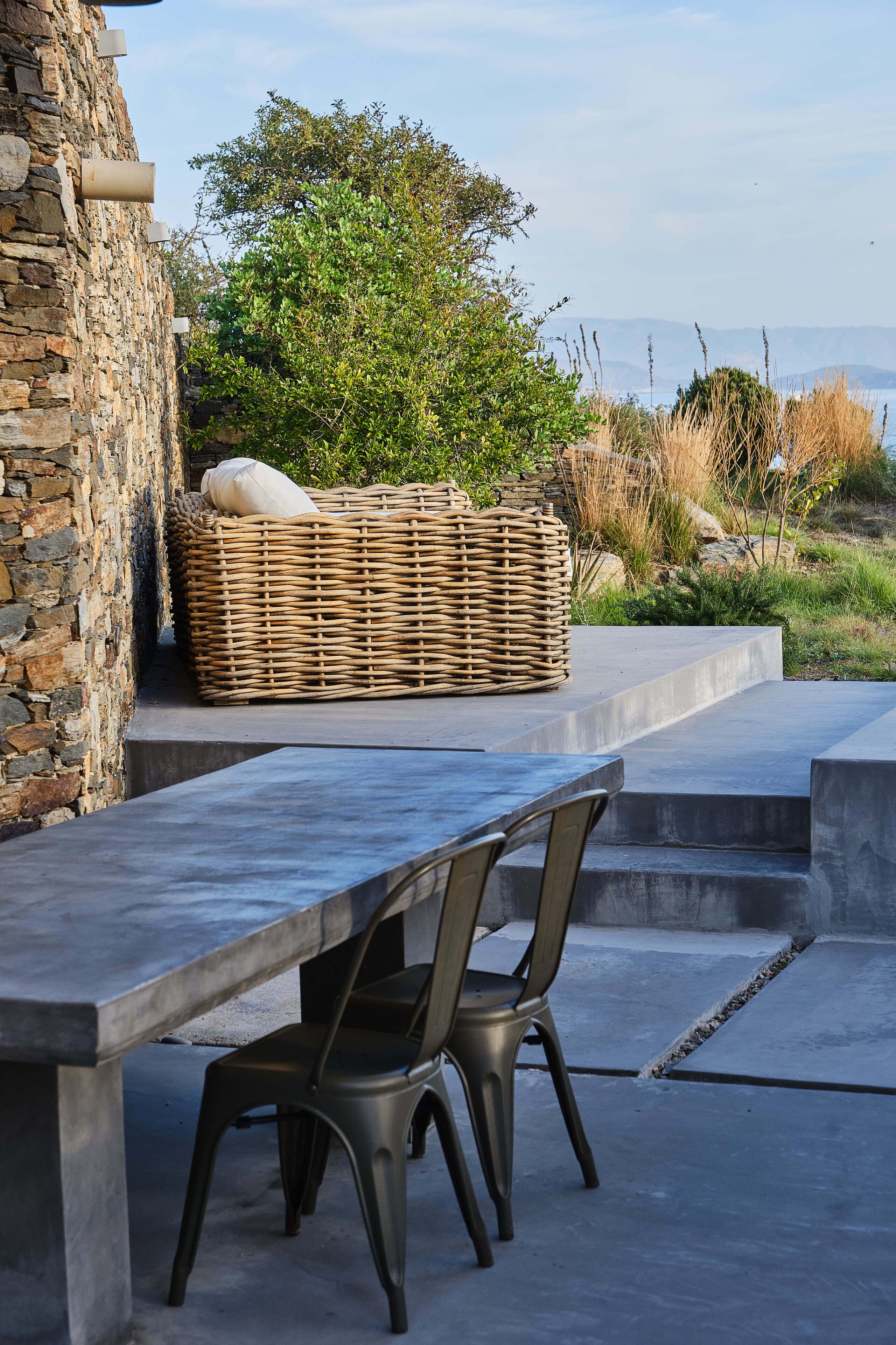 A stone wall, a wicker basket, a concrete table, and a metal chair in an outdoor setting with greenery and a distant view.
