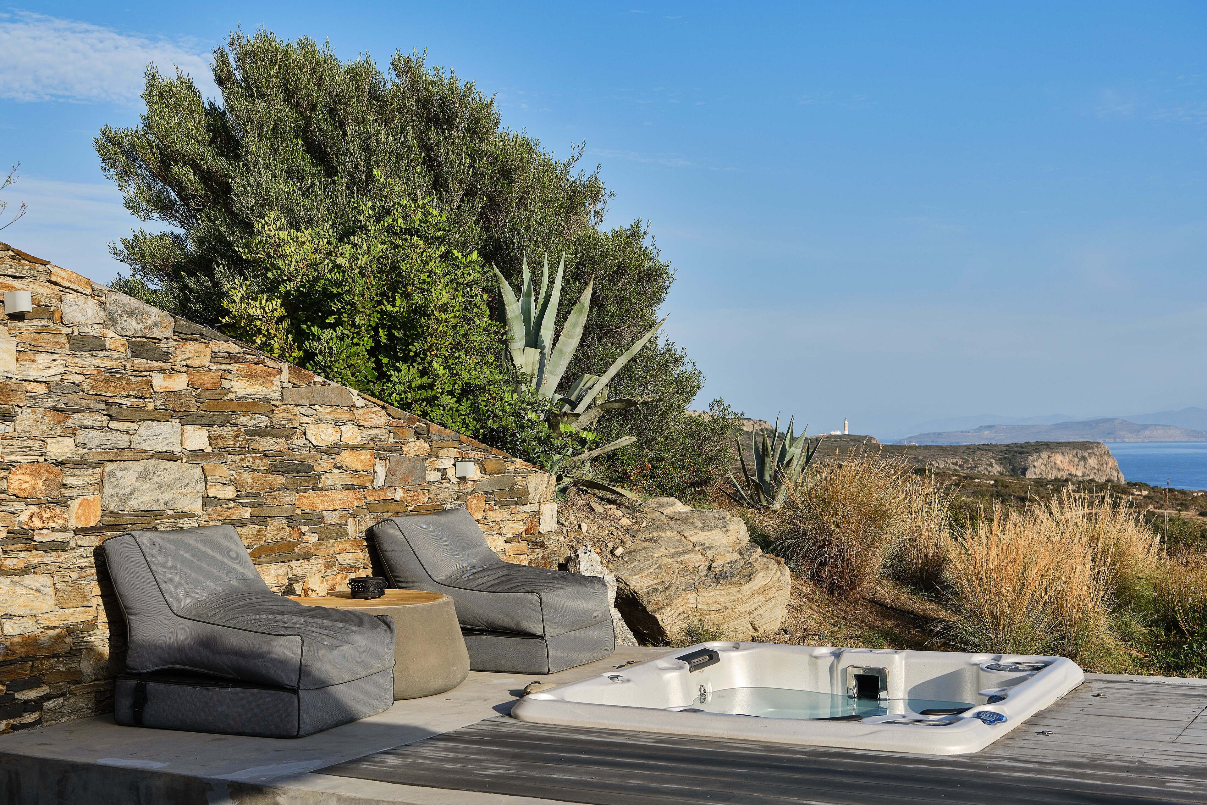 An outdoor seating area with stone walls, two grey lounge chairs, a hot tub, and a view of the sea and cliffs.