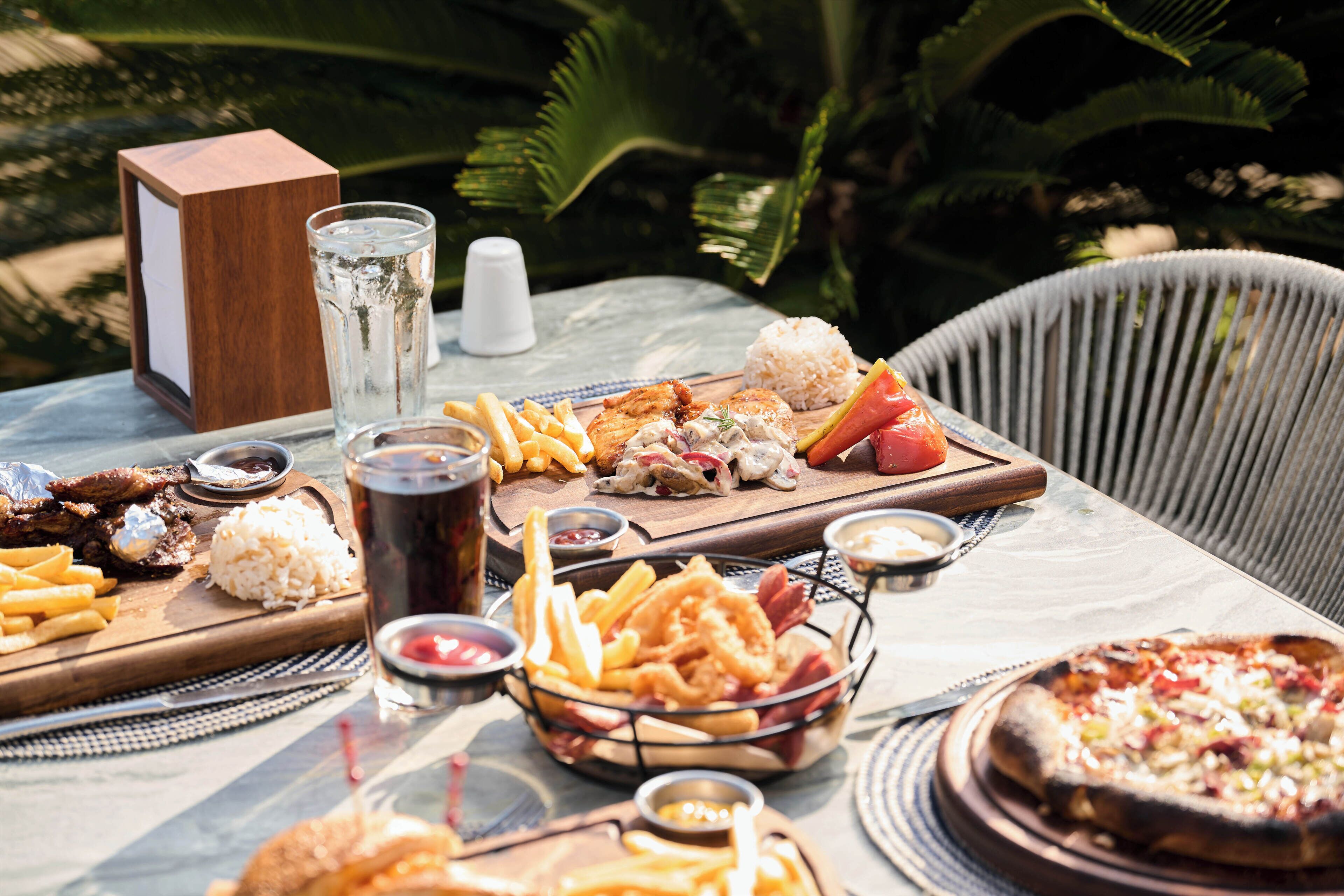 Una mesa dispuesta con diversos alimentos, como pizza, arroz y papas fritas, acompañada de salsas y bebidas.