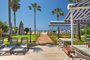 A pathway leading to outdoor seating with umbrellas, palm trees, and a view of the sea.