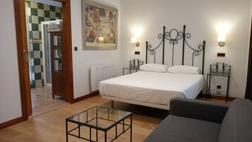 A bedroom with a bed, a glass-top table, a grey sofa, a radiator, a wall-mounted artwork, and a door leading to a bathroom with checkered tiles.