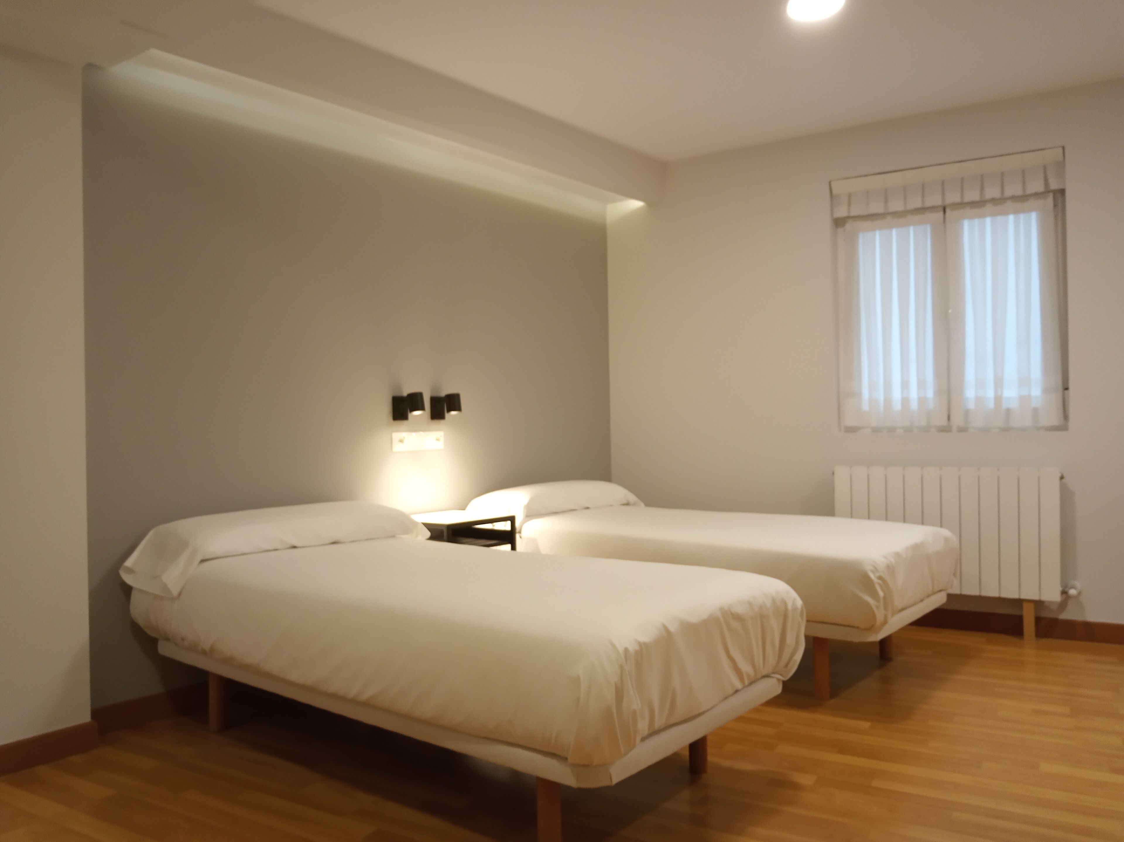 A room with two single beds, a nightstand, a wall-mounted light, a radiator, and a window with curtains.