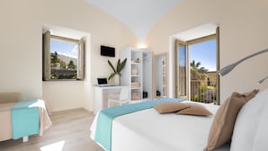 Une chambre avec un lit, un bureau, une chaise et un téléviseur. Il y a une fenêtre qui donne sur des arbres et un immeuble.