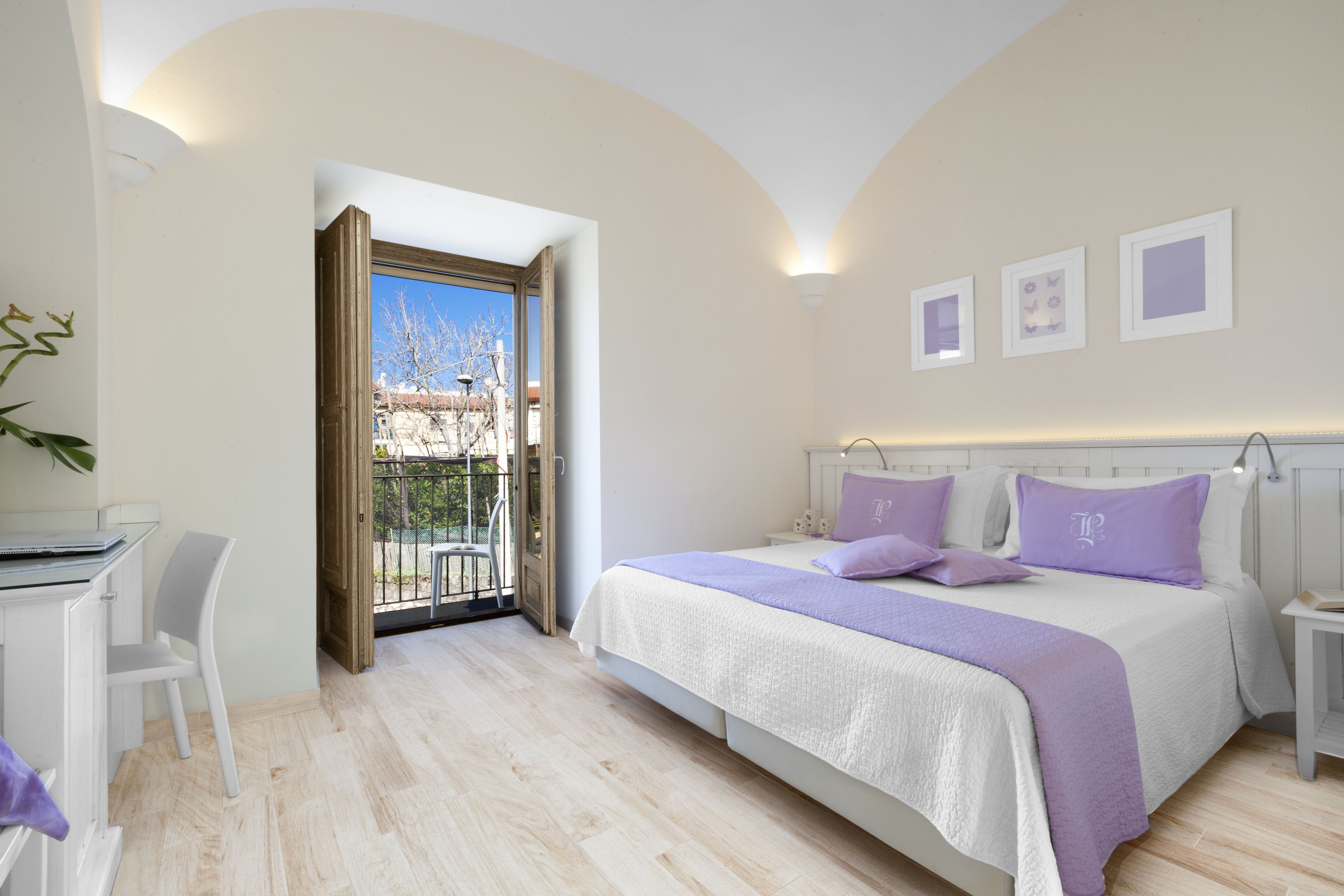 A bedroom with a bed, a desk, and a chair. There is a balcony with a view of the sky and trees.