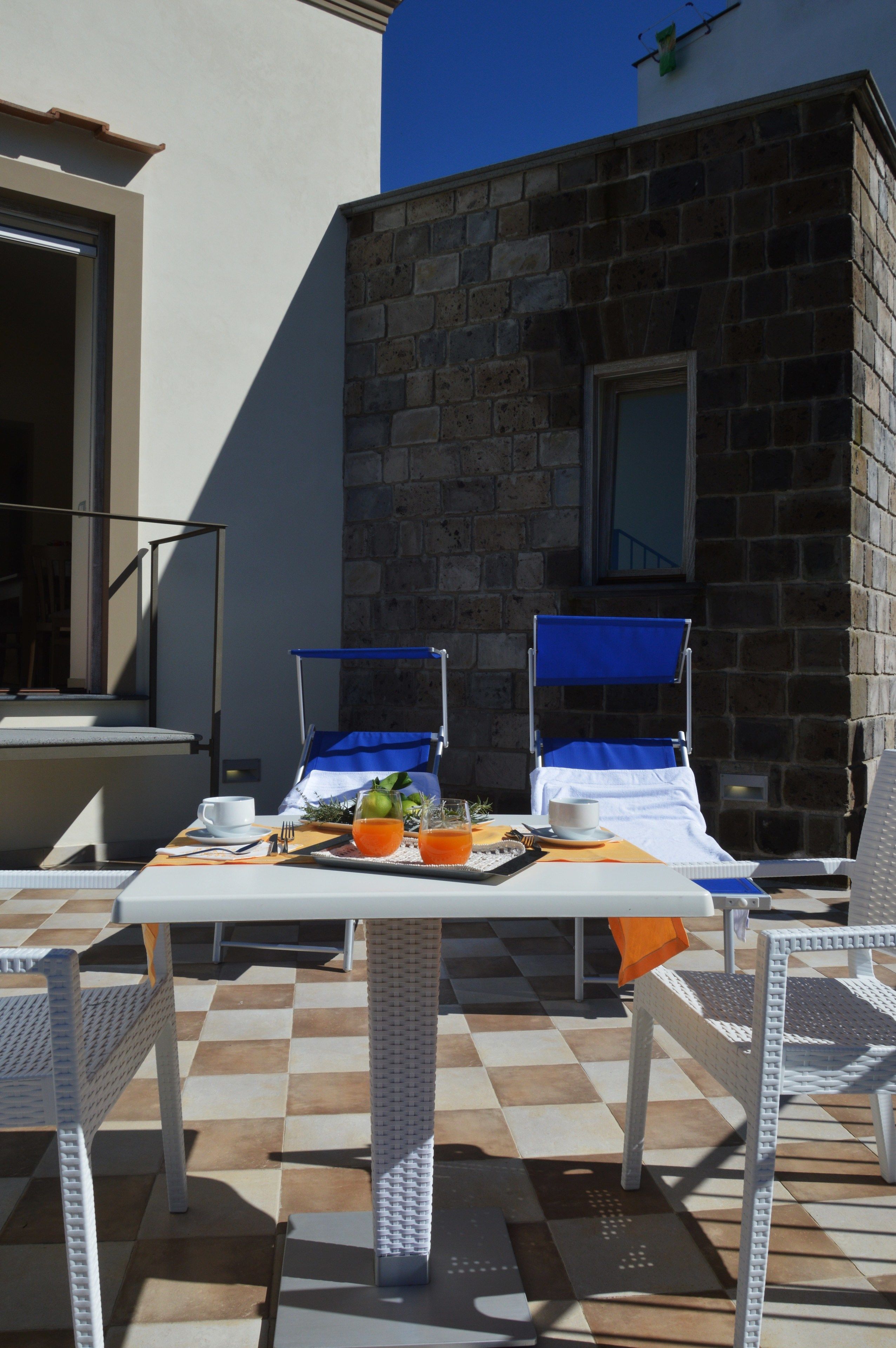 A terrace with a checkered floor, two lounge chairs, a table with breakfast items, and a stone building in the background.