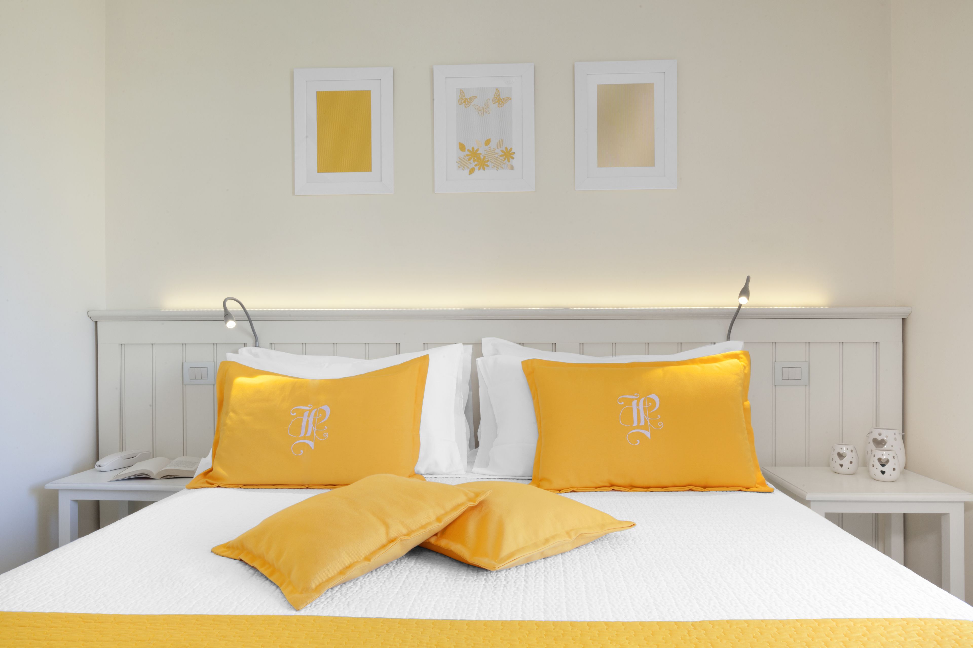 A neatly made bed with white and yellow bedding, a bedside table with a phone and decorative items, and three framed pictures on the wall.