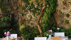 Un espace de restauration en plein air avec des chaises et des tables blanches, entouré de verdure et d’un mur de pierres apparentes.
