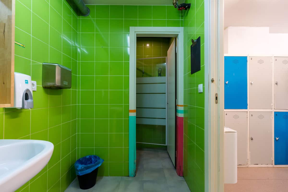 Un baño con azulejos verdes, un lavamanos blanco, un cesto de basura y un conjunto de taquillas.