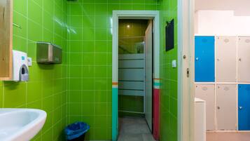 Un baño con azulejos verdes, un lavamanos blanco, un cesto de basura y un conjunto de taquillas.