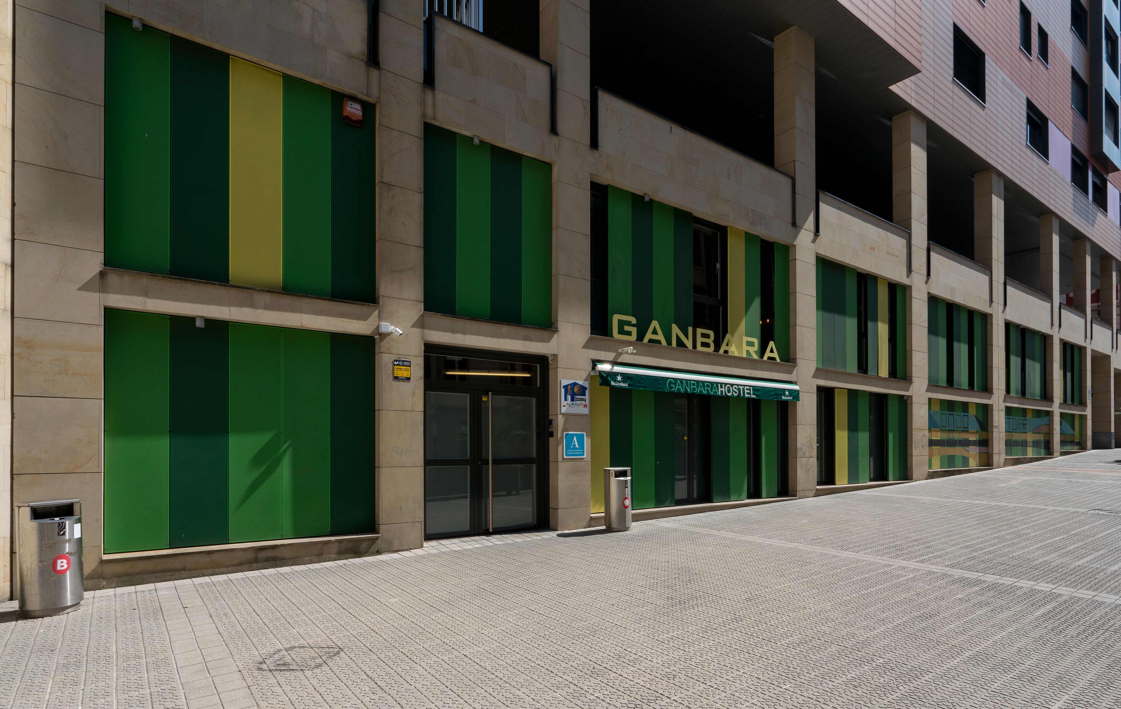 Un edificio con fachada verde y amarilla, un letrero que dice 'GANBARA HOSTEL' y una entrada accesible para sillas de ruedas.