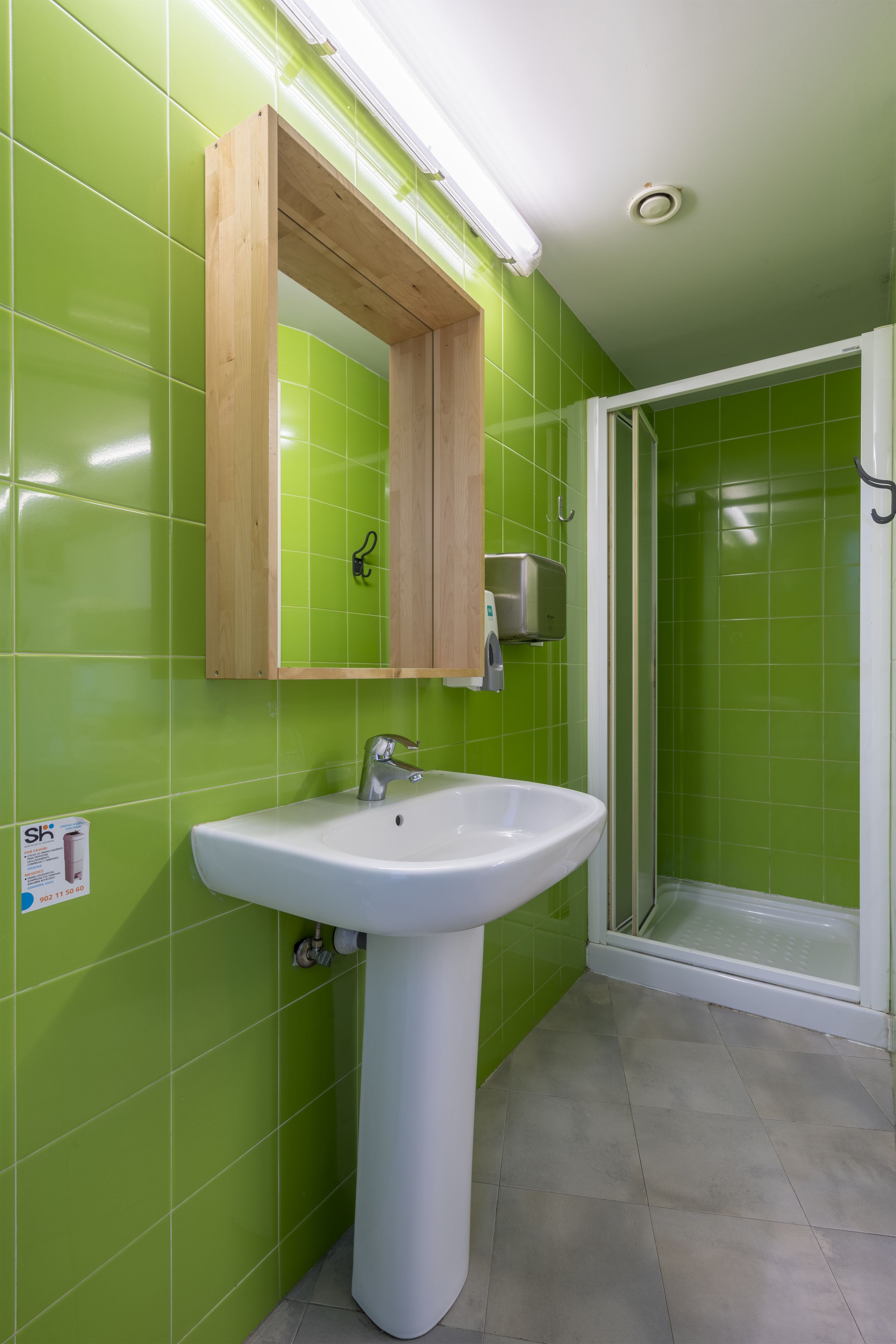 Un baño con un lavamanos blanco, paredes con azulejos verdes y un armario de madera arriba.