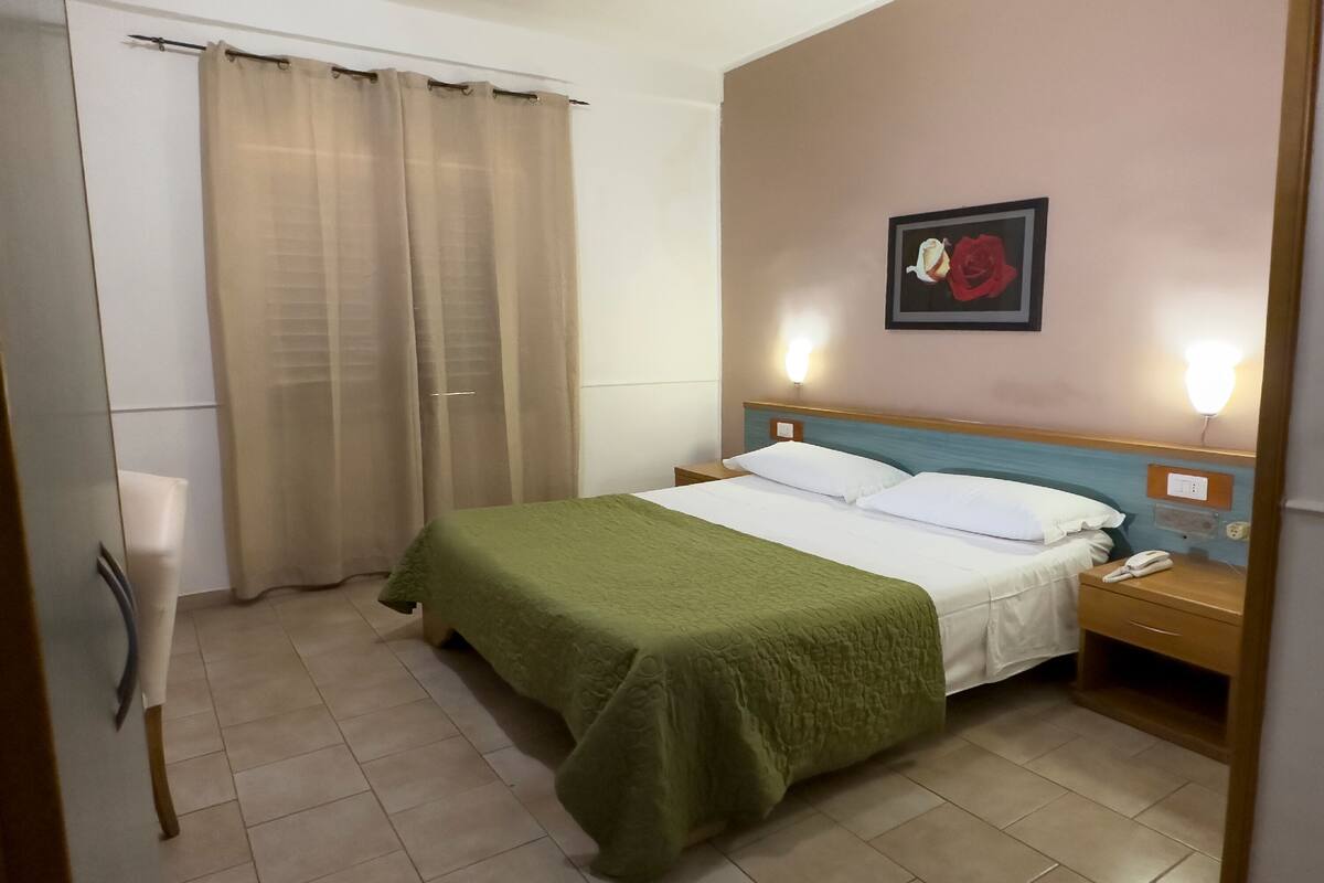 A hotel room with a bed, a nightstand, a wall-mounted phone, and a painting of a rose.