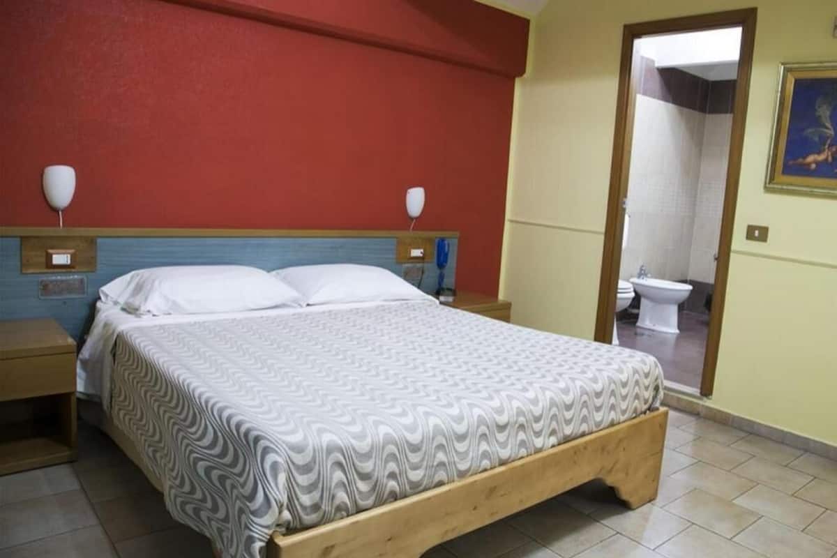 A room with a bed, a nightstand, a wooden headboard, a wall-mounted lamp, and a bathroom visible through an open door.