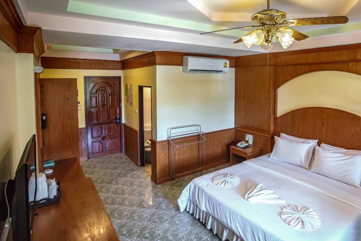 A hotel room with a wooden headboard, a bed with white bedding, a ceiling fan, an air conditioning unit, a bedside table with a lamp, and a bathroom visible through an open door.
