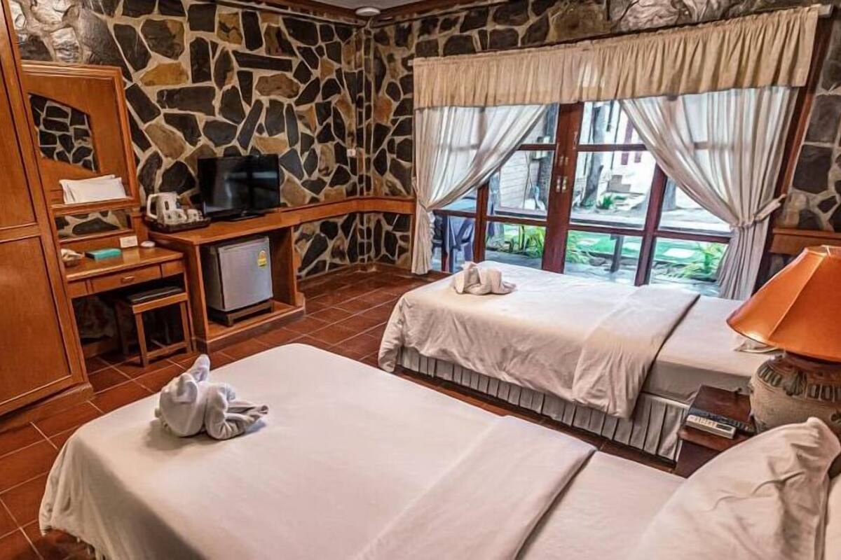A hotel room with two beds, a desk with a television, a stone wall, and a view of greenery.